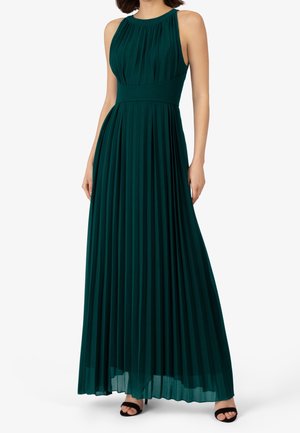 Robe longue plissée sans manches de couleur vert foncé avec taille cintrée, encolure ronde, portée avec des escarpins noirs à bouts ouverts.