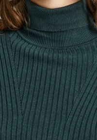 Dunkelgrüner, gerippter Strickpulli mit Rollkragen getragen, der detaillierte vertikale und Zickzackstichmuster auf dem Stoff zeigt.