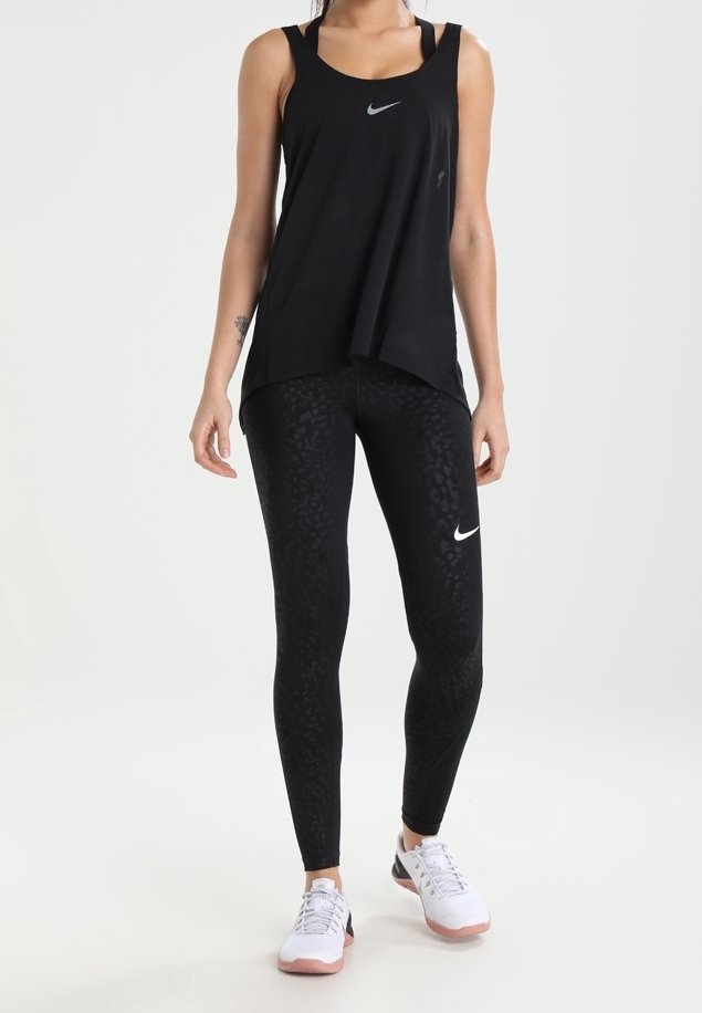 Femme portant un débardeur noir Nike et des leggings à imprimé léopard avec des baskets blanches Nike, debout devant un fond blanc.