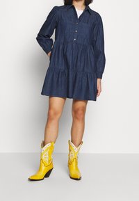 Denim-mekko, jossa on pitkät hihat ja napitusyksityiskohta, yhdistettynä keltaisiin cowboy-kengiin, joissa on valkoisia aksentteja ja kärkikohdat.