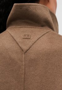 Abrigo de lana marrón visto desde atrás, con un cuello elevado y un sutil detalle de logotipo bordado cerca del escote en una persona con cabello oscuro.