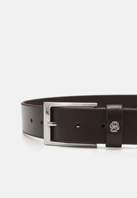 Brun läderbälte med en slät yta, silvermetallspänne och en logotypaccent. Har justerbara hålplatser för storlek.