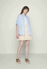 Camisa azul clara de mangas curtas com botões por cima de um top branco, combinada com uma saia plissada floral em tons pastel e sandálias rasas castanhas.