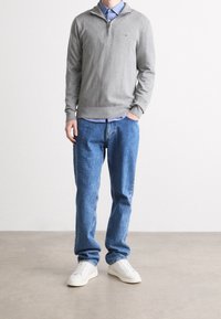 Maglione grigio con zip e collo alto sopra una camicia azzurro chiaro, abbinato a jeans blu larghi e sneaker bianche, con un design semplice e casual.