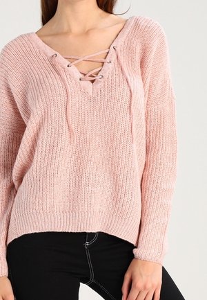 Femme portant un pull en maille rose clair avec un col en V lacé et des manches longues, associé à un pantalon noir avec des coutures blanches.