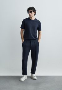 Camisa de manga curta texturada azul-marinho combinada com calças azul-marinho feitas à medida. Modelo usa óculos de sol e ténis brancos. Design minimalista.