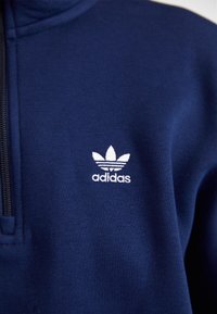 Tamsiai mėlynas užtrauktuku užsegamas džemperis, pagamintas iš minkštos medžiagos, su baltu „Adidas“ logotipu ir trimis dryželiais ant krūtinės.