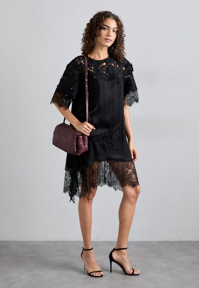 Μαύρο φόρεμα με δαντέλα, περιθώρια σε σχήμα κυματοειδούς και κοντά μανίκια. Το μοντέλο κρατάει μια μπορντό τσάντα και φοράει μαύρες γόβες με λουράκια.