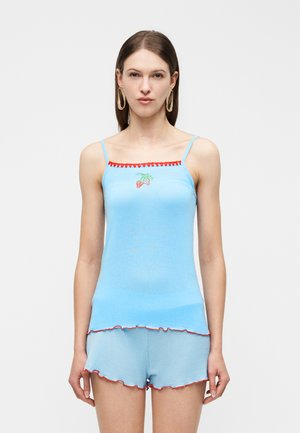 Nő világoskék, piros szegéllyel és eperhímzéssel díszített camisole és rövidnadrág szettben, egy egyszínű háttér előtt áll.