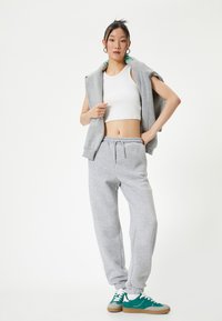 Grijze katoenen joggingbroek en mouwloze hoodie over een witte crop top. Model met teal en grijze sneakers, staand tegen een witte achtergrond.