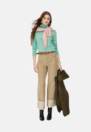 Giovane donna con maglione turchese e sciarpa rosa, pantaloni beige con risvolto, stivali neri, tiene in mano una giacca imbottita scura.