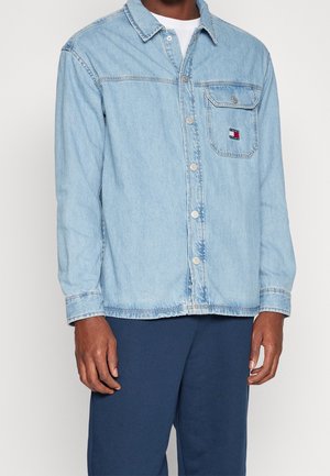 Homme portant une chemise en denim bleu clair à boutons avec une poche poitrine présentant un petit logo rectangulaire rouge, blanc et bleu.