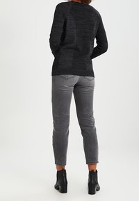 Černý pletený svetr s texturovaným designem, v kombinaci se šedými džíny. Model má na sobě černé kotníkové boty. Pohled zezadu, ukazující střih a siluetu.