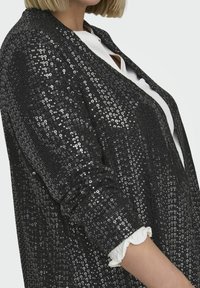 Mustmustel põhja ja pehme disainiga must sädelev bleiser, millel on lühikesed varrukad, tekstureeritud pind ja volangiline valge aksent varruka servas.