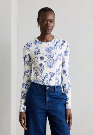 Femme portant un haut blanc à manches longues avec des motifs bleus complexes de soleil, fleurs et animaux, associé à un jean bleu foncé taille haute.