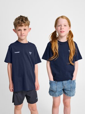 To børn står side om side iført mørkeblå Hummel t-shirts; dreng i sorte shorts, pige med rødt hår i denimshorts med hænderne i lommerne.