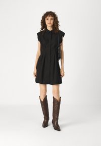 Μαύρο φόρεμα κοντό μανίκι με γιακά και εμπρός πτυχές, φορεμένο με καφέ μοτίβο καουμπόικες μπότες. Το φόρεμα έχει χαλαρή γραμμή και μήκος πάνω από το γόνατο.