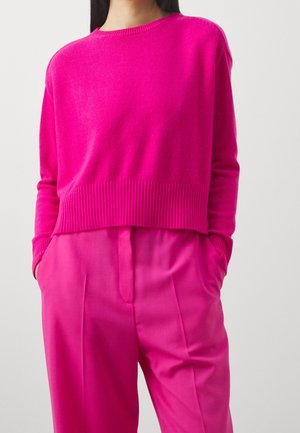 Femme portant un pull à manches longues rose vif rentré dans un pantalon rose taille haute assorti, les mains dans les poches, sur fond uni.