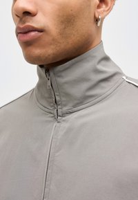 Suure plaan inimesest, kes kannab helehalli lukuga jope kõrge kraega ja hõbedast rõngakujulist kõrvarõngast vasakus kõrvas.