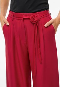 Rode wijde pantalon met een gladde textuur, voorzien van een stoffen roosdetail op de riem en zijzakken. Draag het met een zwarte top.