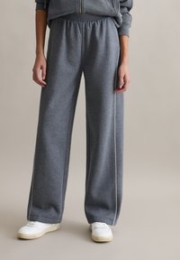 Szare szerokie spodnie dresowe z miękkiego materiału o gładkiej fakturze i białym paskiem po bokach. W połączeniu z białymi trampkami.