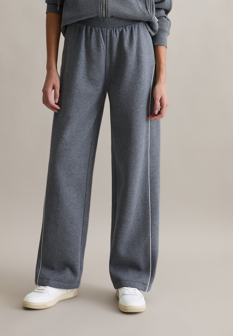 Szare szerokie spodnie dresowe z miękkiego materiału o gładkiej fakturze i białym paskiem po bokach. W połączeniu z białymi trampkami.