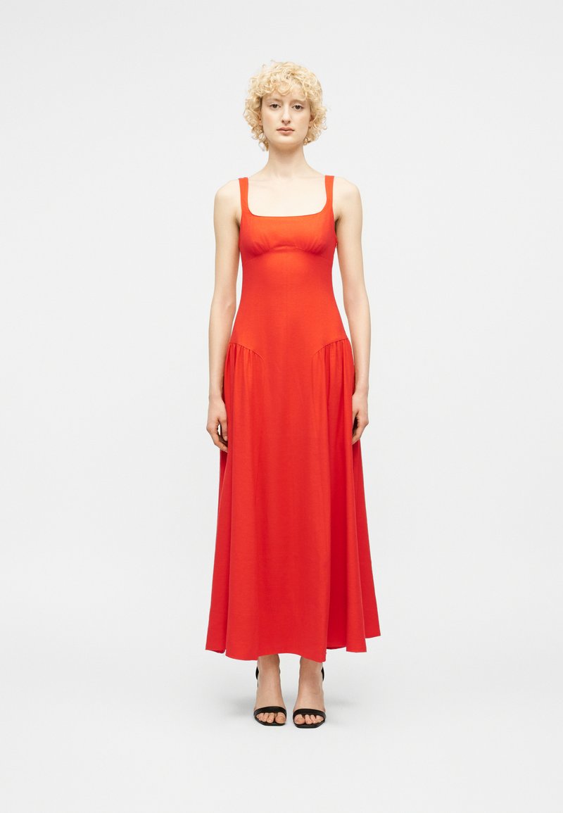 Γυναίκα με ξανθά σγουρά μαλλιά, ντυμένη με ένα μακρύ κόκκινο φόρεμα χωρίς μανίκια και μαύρα ανοιχτά πέδιλα, στέκεται μπροστά σε απλό λευκό φόντο.