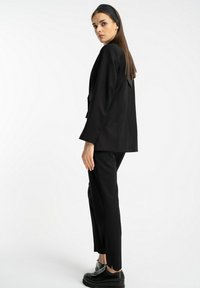 Czarna marynarka o dopasowanym kroju z zapięciem na jeden guzik, w zestawie z dopasowanymi spodniami wąskimi. Gładka tkanina, minimalne detale oraz płaskie czarne buty.