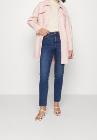 Lång jacka i rosa ullblandning med stora fickor, kombinerad med åtsittande mörkblå jeans och vita klacksandaler. Slät textur och enkel design.