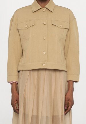 Giacca corta beige con chiusura a bottoni e tasche con patta, indossata sopra una gonna plissettata beige chiaro.