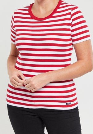 Osoba ubrana w koszulkę z krótkim rękawem w czerwono-białe poziome paski z czerwonym kołnierzem, w czarnych spodniach, z rękami splecionymi na wysokości pasa.