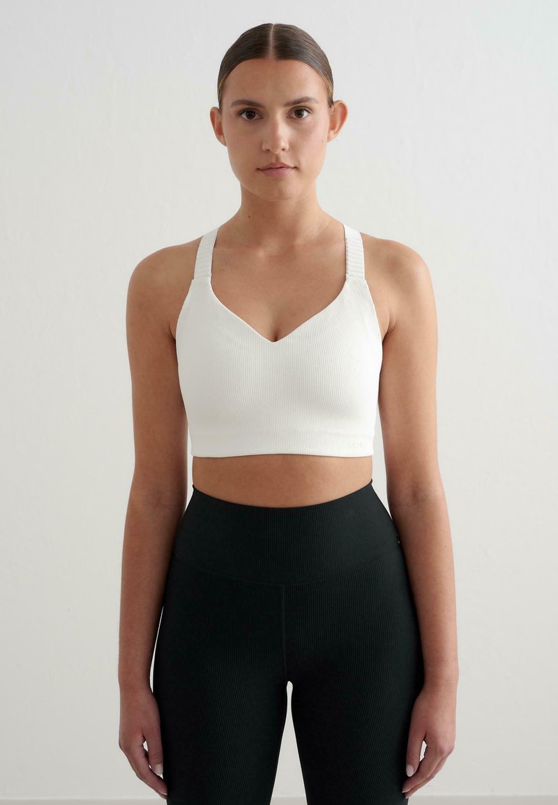 Biely rebrovaný crop top s výstrihom do V a širokými ramienkami. Skombinovaný s čiernymi rebrovanými legínami s vysokým pásom. Hladká textúra, priliehavý dizajn.