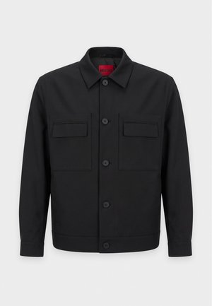 Homme portant une casquette noire, une veste noire sur un t-shirt graphique, un jean foncé, une ceinture noire et des mocassins noirs, debout les mains dans les poches.