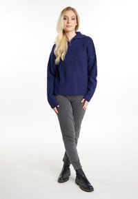 Marineblauwe geribbelde trui met een ritskraag, lange mouwen en een ontspannen pasvorm, gecombineerd met grijze slim-fit broek en zwarte enkellaarsjes.