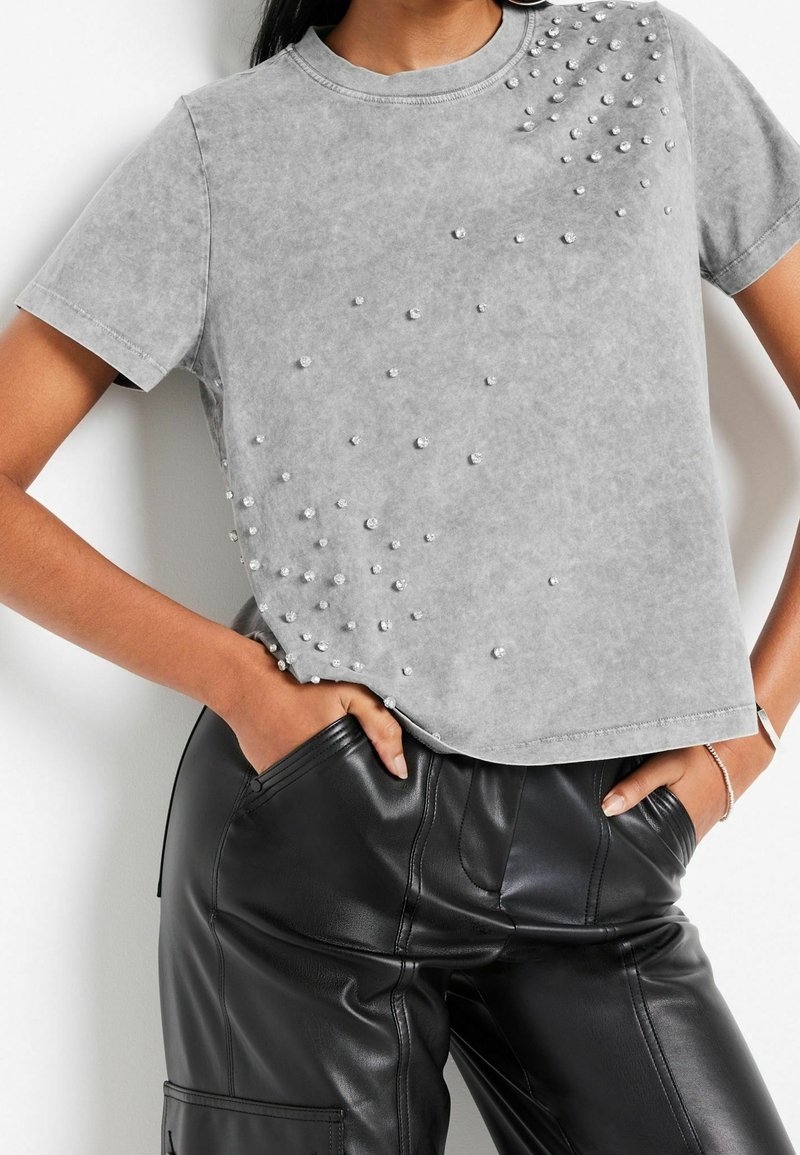 Personne portant un t-shirt gris à manches courtes avec des ornements en perles éparpillées et un pantalon en cuir noir, les mains dans les poches.