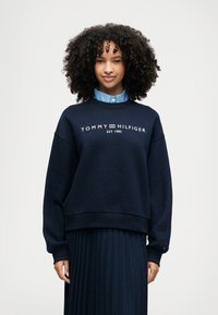 Ναυτικό φούτερ με λευκό λογότυπο "TOMMY HILFIGER EST. 1985", ριμπ λαιμόκοψη και μανσέτες; φορεμένο πάνω από ένα ανοιχτό μπλε πουκάμισο με γιακά και πλισέ φούστα.
