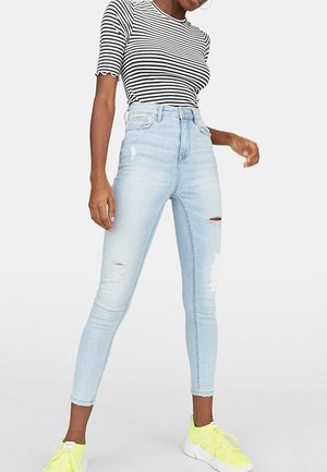 Mujer vestida con jeans skinny rasgados de color azul claro, camiseta de rayas en blanco y negro de manga corta, y zapatillas de deporte amarillas brillantes, de pie sobre un fondo blanco liso.