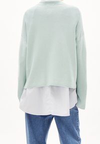 Pull court vert menthe clair avec une finition texturée, superposé à un ourlet de chemise blanche. Coupe décontractée avec épaules tombantes et poignets côtelés.