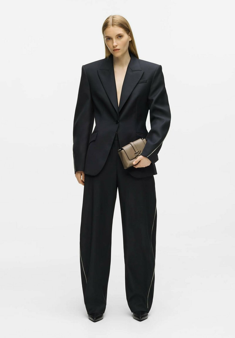 Femme aux longs cheveux blonds porte un blazer noir oversize et un pantalon large, tenant un petit sac taupe sur fond blanc.