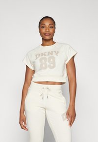 Kreemas cropped džemperis ar "DKNY 89" bēša krāsā, savienots ar atbilstošām bēšām sporta biksēm ar logo akcentu un aukliņu jostasvietā.