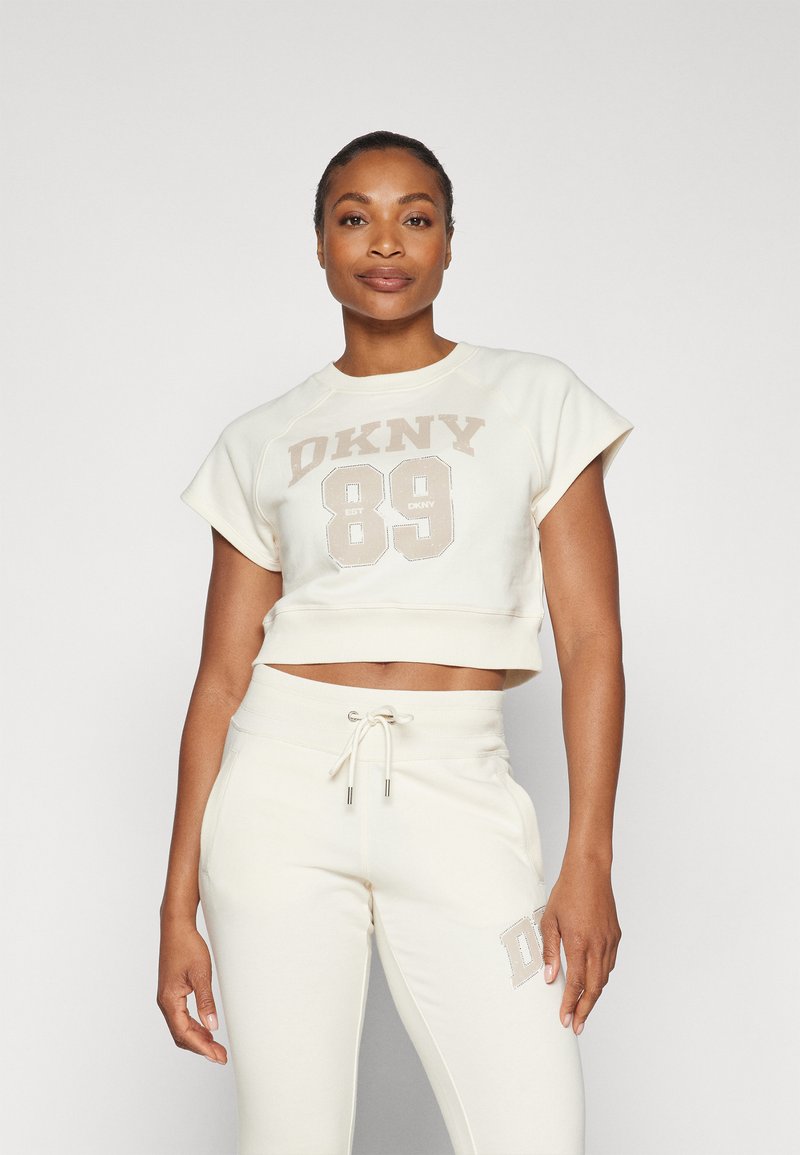 Kreemas cropped džemperis ar "DKNY 89" bēša krāsā, savienots ar atbilstošām bēšām sporta biksēm ar logo akcentu un aukliņu jostasvietā.