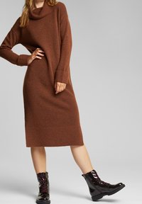 Vestito a maglia marrone con collo ad anello, polsini e orlo a coste. Modella che indossa stivali neri lucidi con lacci e dettagli bordeaux.