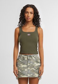 Oliwkowy crop top o kwadratowym dekolcie z różowym haftowanym logo, w zestawie z szarym spódnico-spodnie w kamuflaż z kieszeniami i bocznymi rozcięciami.
