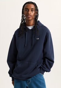 Felpa blu navy realizzata in cotone morbido, con tasca frontale, cappuccio con lacci e piccolo logo sul petto. Indossata con jeans blu.