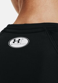 Czarna koszulka sportowa o gładkiej strukturze, z okrągłym białym logo na plecach, zawierającym symbol Under Armour oraz miejsce na imię.