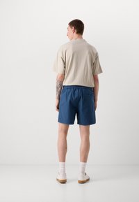 Chemise beige à manches courtes, shorts bleus, taille élastique, détail de poche arrière. Chaussures décontractées avec semelles claires et chaussettes blanches à côtes.