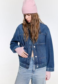 Jachetă din denim de culoare albastră închisă, cu o croială lejeră, având nasturi argintii și manșete tricotate roz. Asortată cu blugi de culoare albastră deschisă.