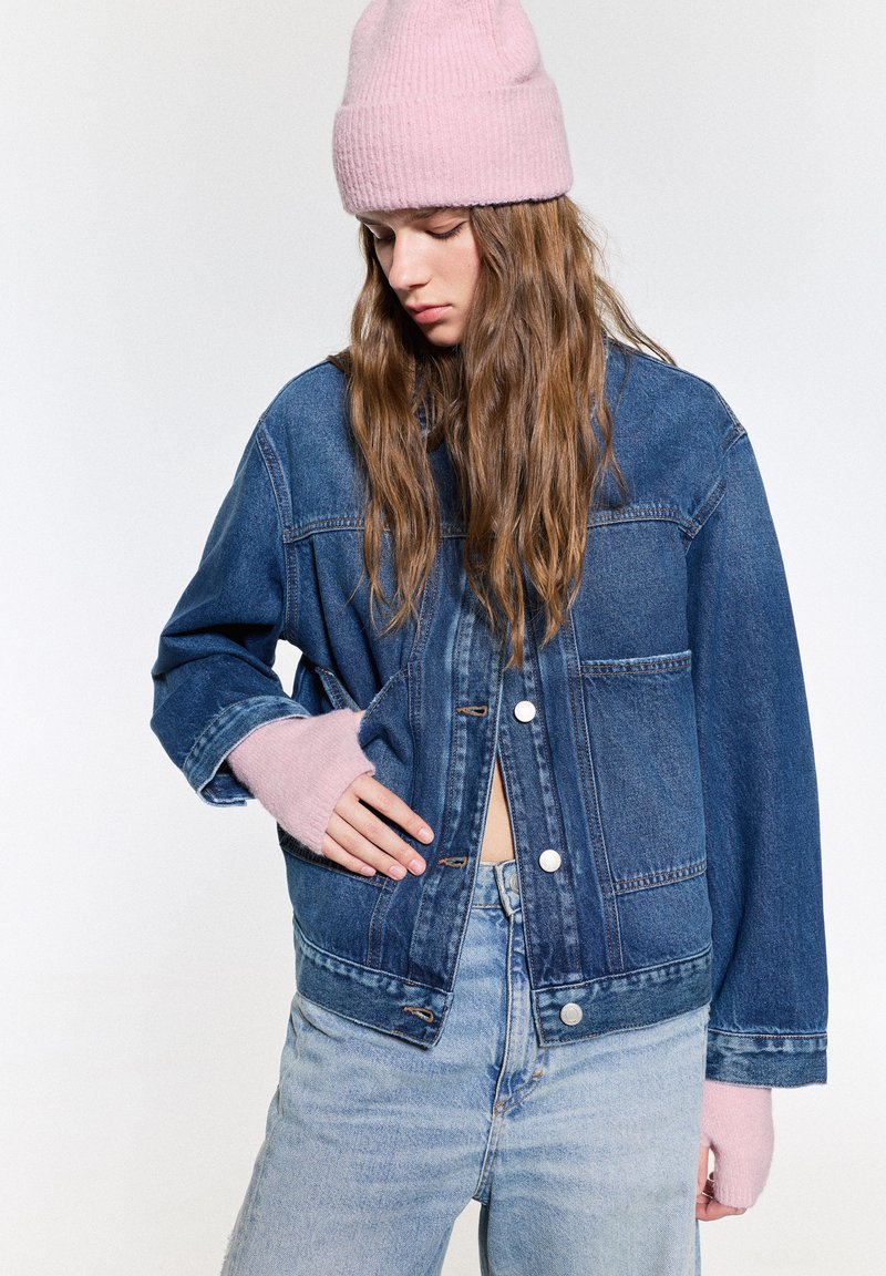 Jachetă din denim de culoare albastră închisă, cu o croială lejeră, având nasturi argintii și manșete tricotate roz. Asortată cu blugi de culoare albastră deschisă.