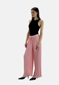 Schwarzes ärmelloses Oberteil kombiniert mit hellrosa, weit geschnittenen, plissierten Hosen. Die Hose hat einen elastischen Bund und einen Kordelzug. Schwarze spitze Schuhe.