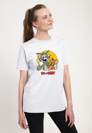 T-shirt bianca in cotone con una grafica colorata di Tom e Jerry su uno sfondo a cerchio giallo. La maglietta ha maniche corte e colletto girocollo.
